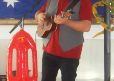 Musician Rob T singing and playing his ukulele standing in front of a large Australian flag