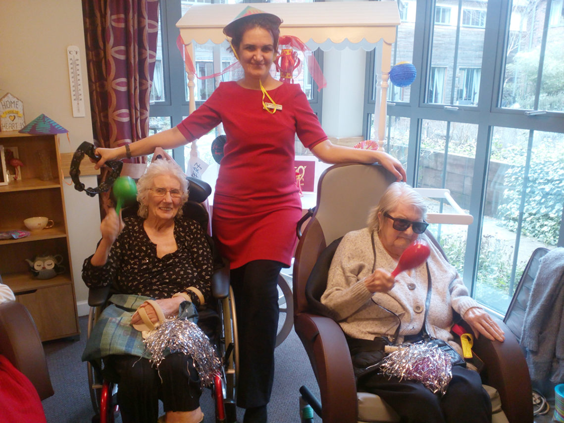 Two seated residents with a staff member between them enjoying musical instruments at Hengist Field Care Home
