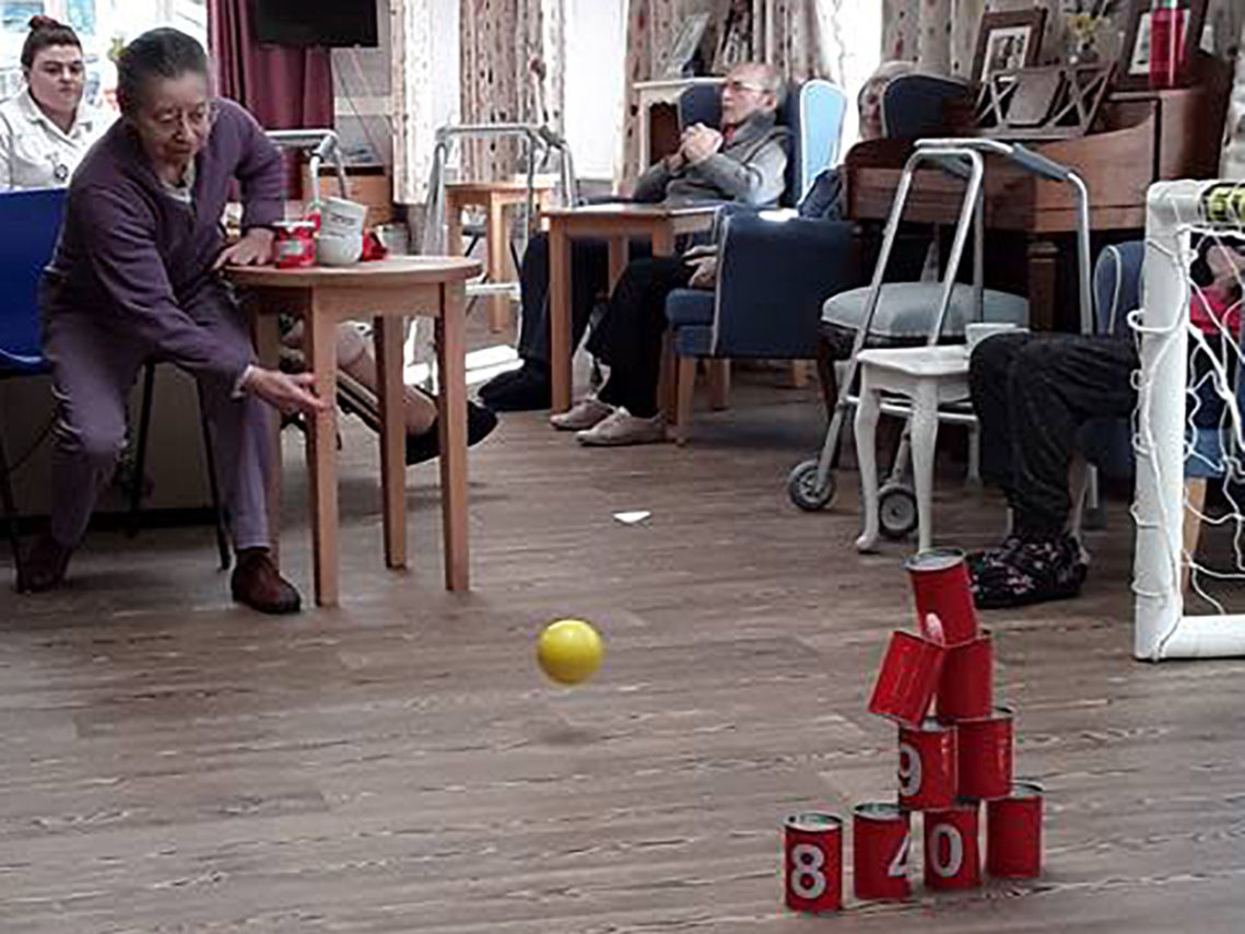 A seated lady resident rolling a ball at a stack of tin cans in the lounge at Abbotsleigh Care Home