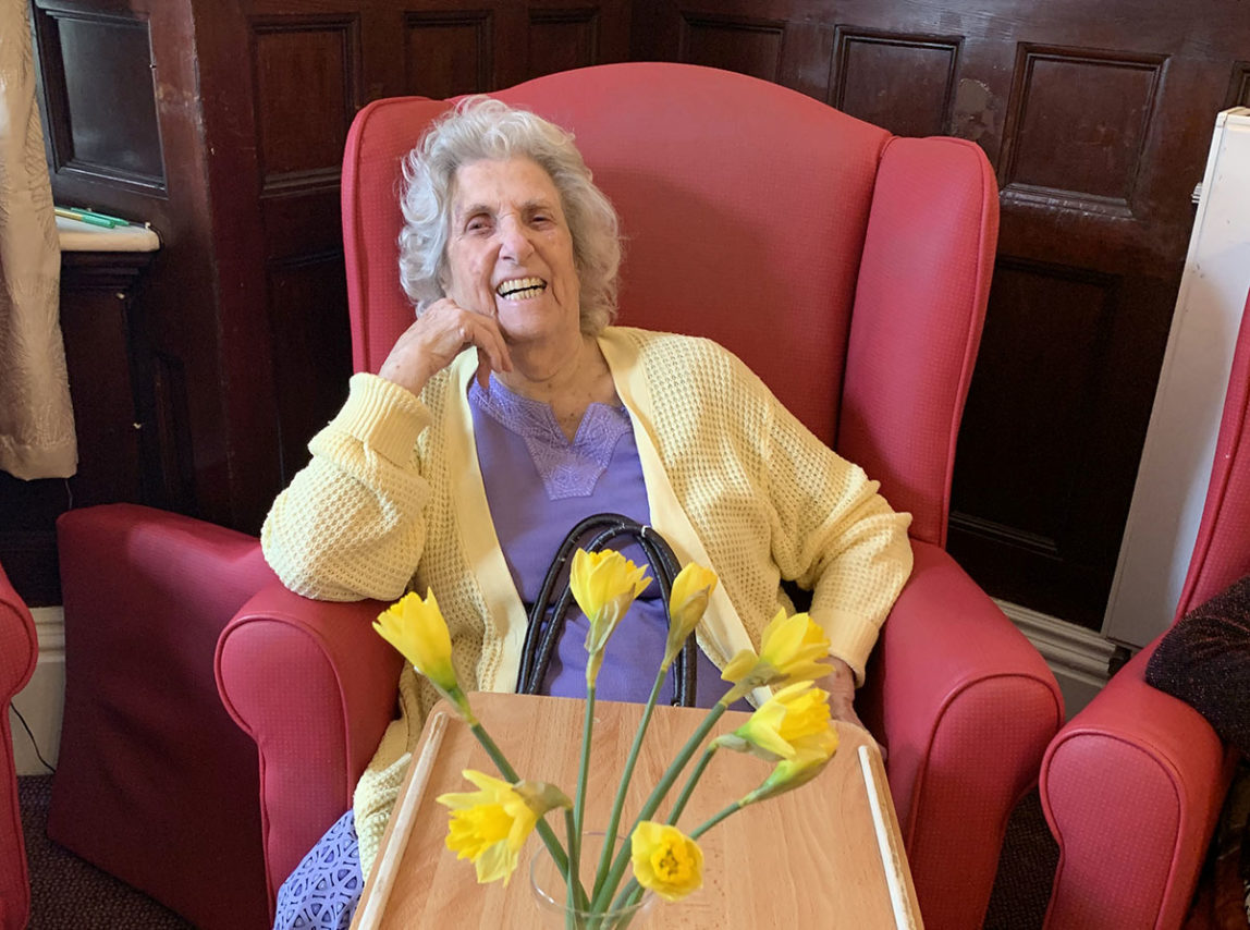 Female resident sitting in a chair smiling to the camera, with a vase of daffodils in front her