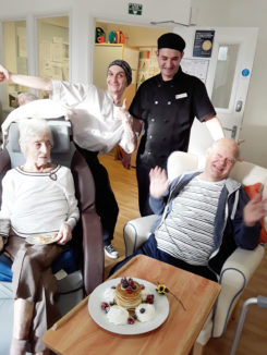 Two residents and two kitchen staff smiling together over their decorated stack of pancakes